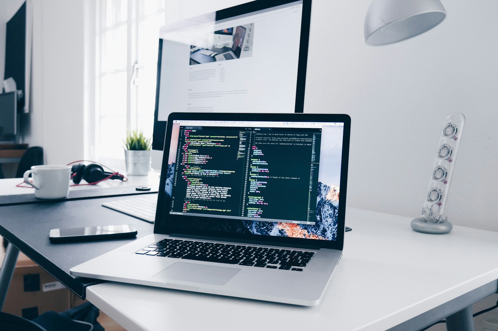 Mesa de trabajo con un portátil abierto en un editor de código y un monitor detrás con una página web.